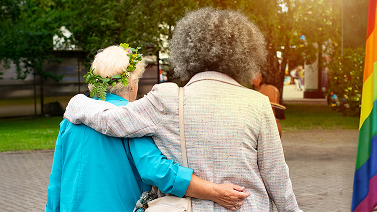 An elderly queer couple walking with arms linked. There is a pride flag next to them.